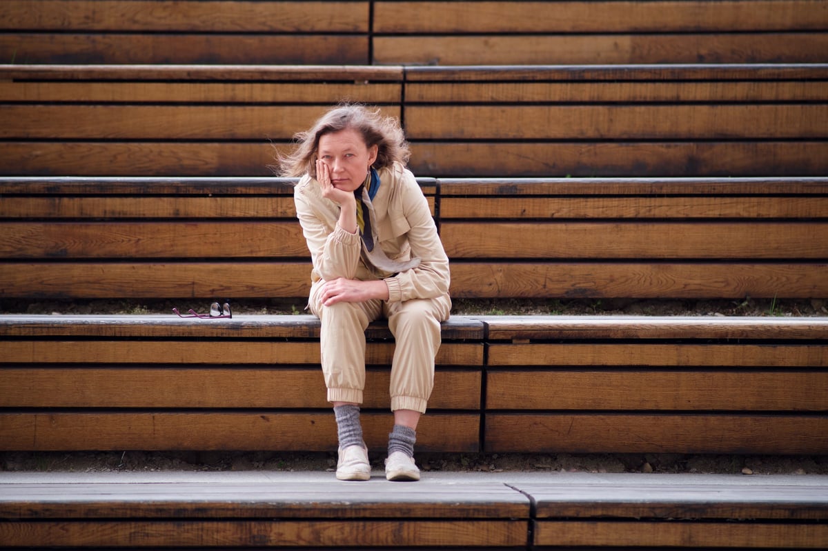 White woman in her 50s sitting alone on a wooden bench, deep in thoughtful reflection and contemplation, embracing midlife renewal and new possibilities