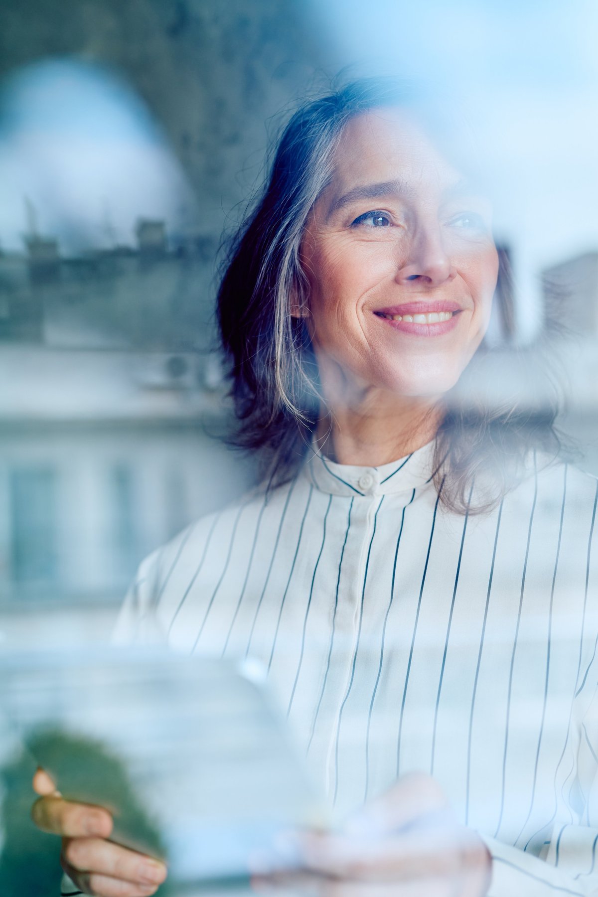 Mature woman smiling, looking out through a window with city reflections, holding a tablet. Reflecting on future plans