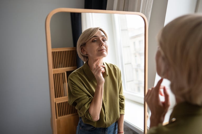 Beautiful positive middle aged woman looking in mirror and checking makeup in reflection, daily ritual of self love and admiring own beauty. Smiling confident female looking satisfied with outlook.