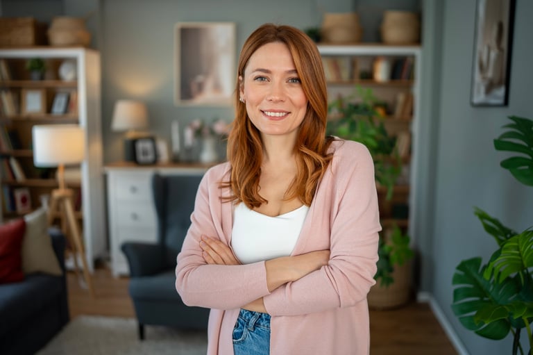 Redhead woman standing in a spacious living room, arms crossed and smiling directly at the viewer, expressing confidence and a relaxed, at-home feeling