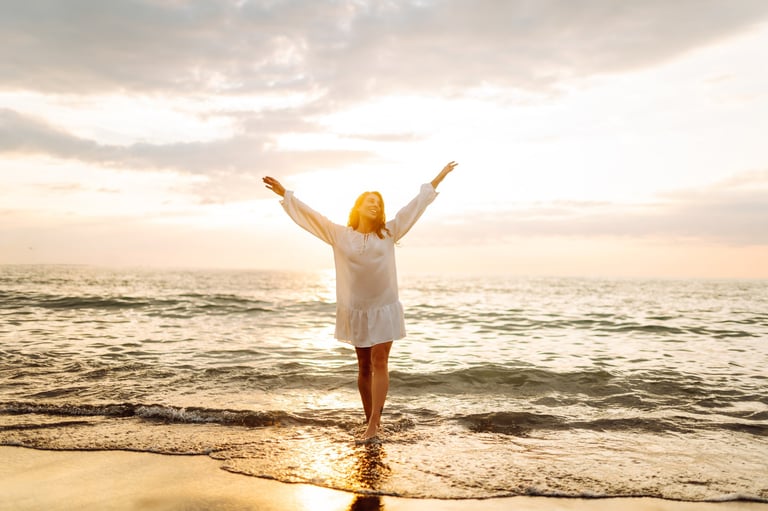 A woman stands by the shore with the ocean behind her, curly hair, laughing as the sun sets creating a warm glow